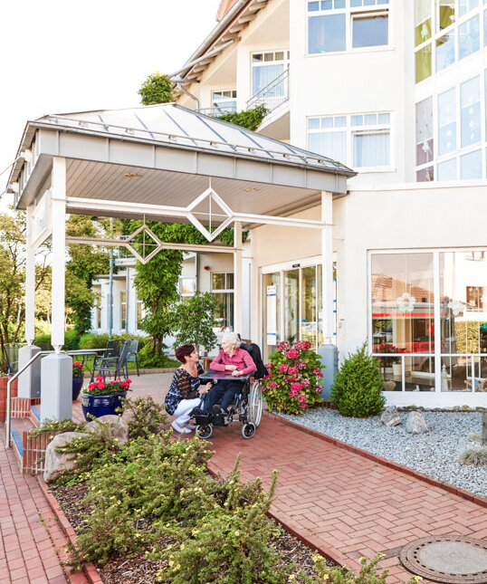 Bewohnerin im Rollstuhl mit Pflegerin vor dem Eingangsbereich des Geriatrie-Zentrum Rhenanus in Bad Sooden